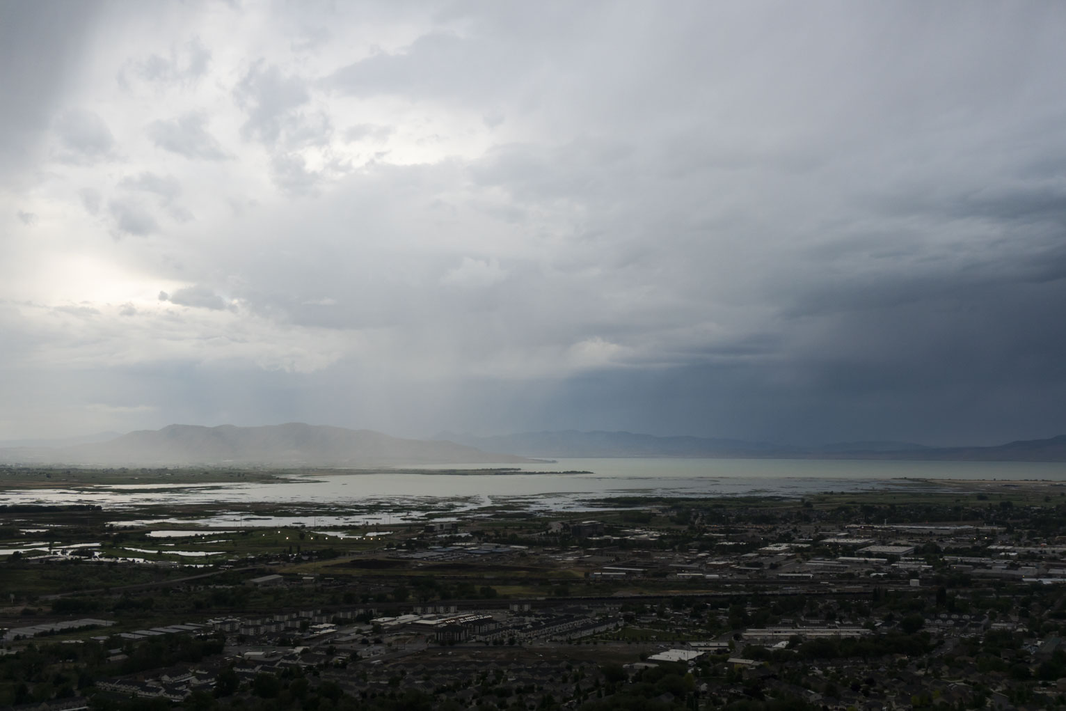 Rain over a lake and mountains with a shadowed town in the foreground, the left side is awash in light from a thinning in the clouds